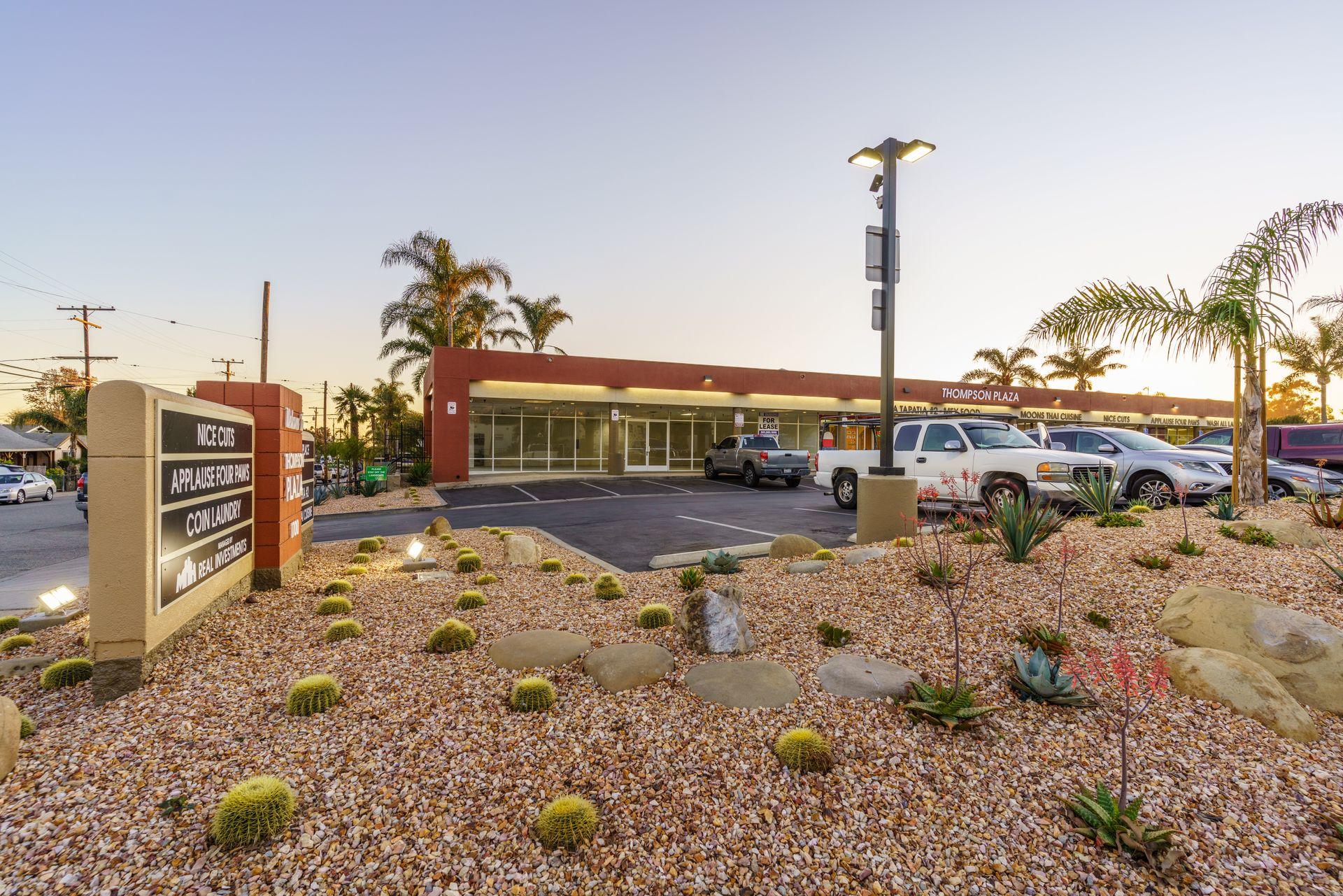 Desert-style shopping plaza with cacti and parked cars at sunset.