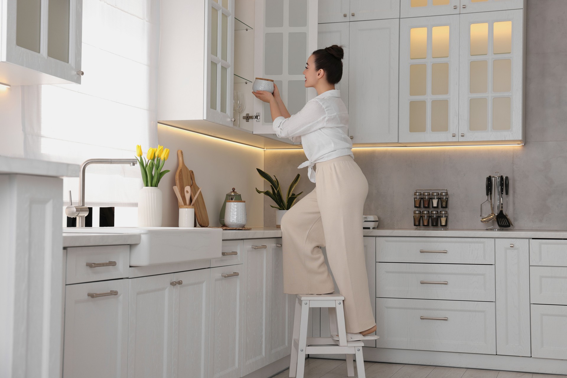 Woman on ladder putting white jar into cupboard at home